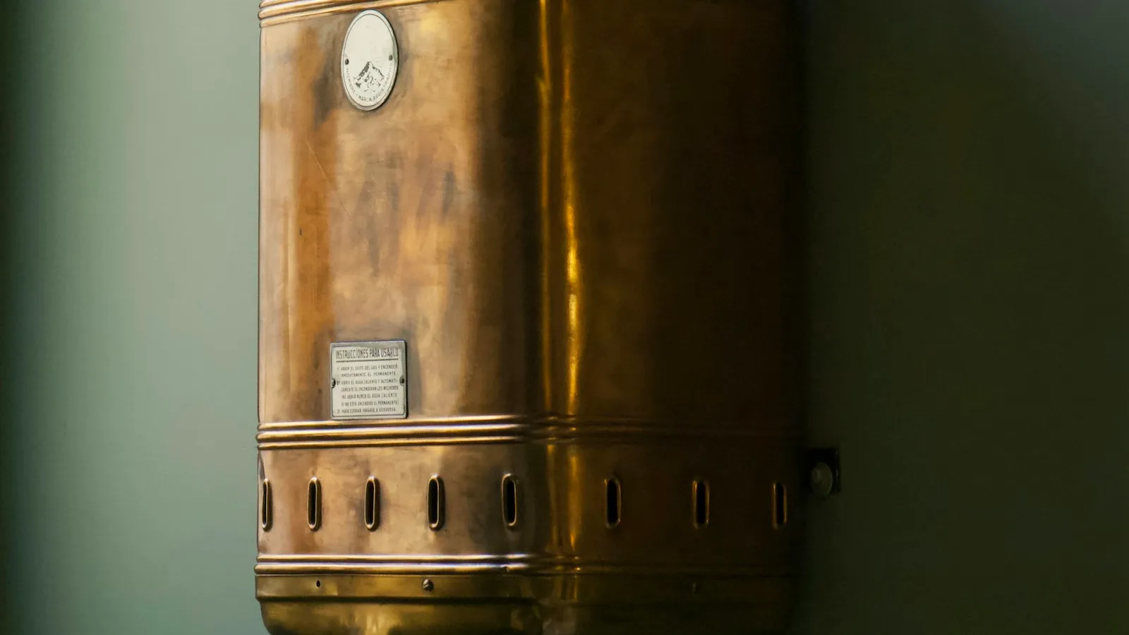 A brass water heater tank with a gauge against a green wall