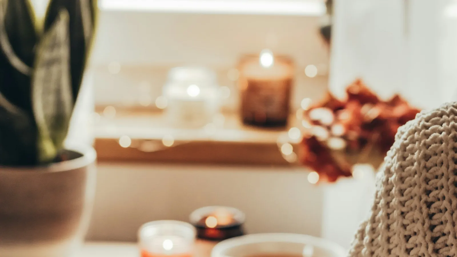 Person in a knit sweater holding a mug near candles and a plant